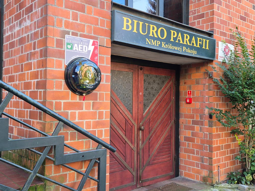 Wejście do budynku parafialnego z drewnianymi drzwiami, obok których zamontowany jest defibrylator AED.