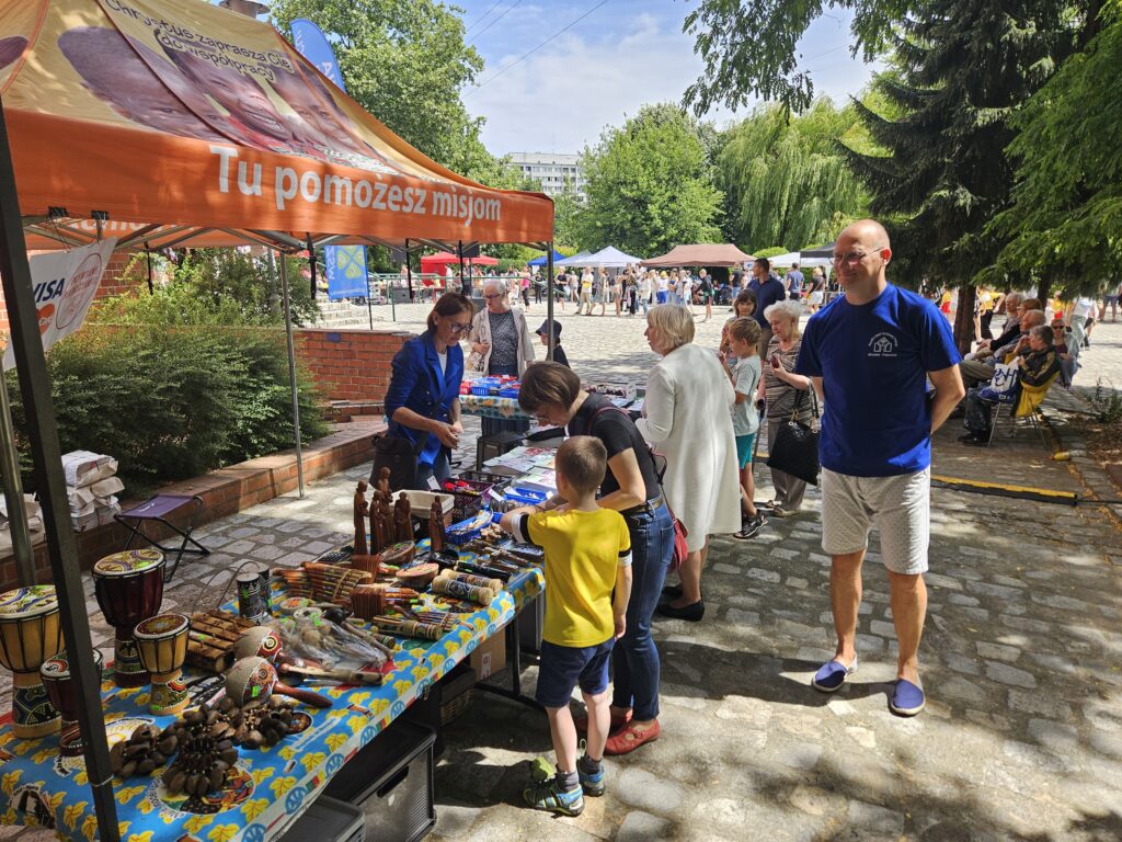 Dzieci i dorośli oglądają stoisko z rękodziełem ustawione pod pomarańczowym namiotem podczas wydarzenia plenerowego.