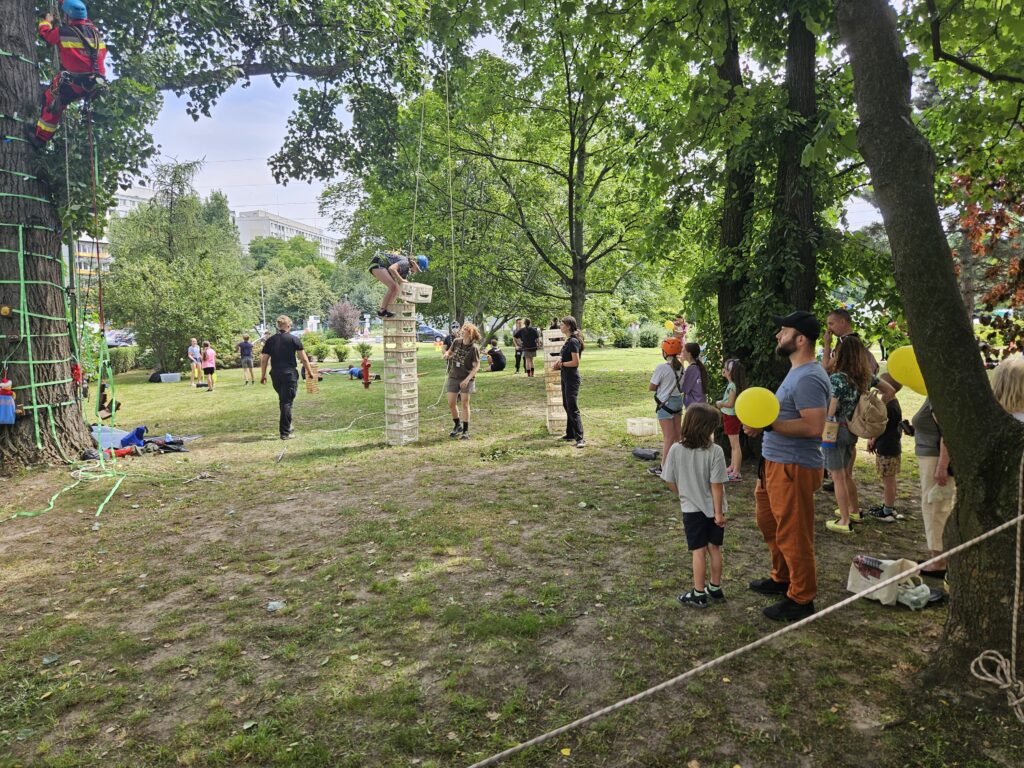 W parku osoba w kasku i uprzęży wspina się po wysokim stosie plastikowych skrzynek, asekurowana linami przez instruktorów; obok inna osoba wspina się na drzewo, a dzieci i dorośli obserwują aktywność, trzymając żółte balony.