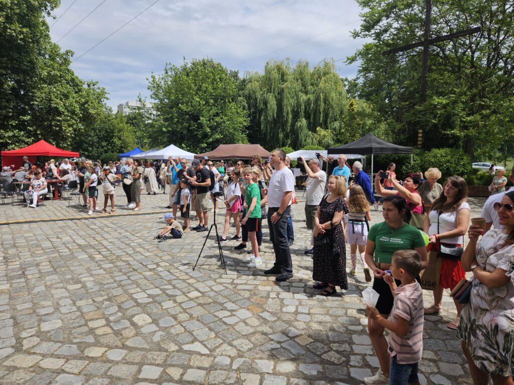 Grupa dorosłych i dzieci stoi na placu podczas festynu, obserwując występ na scenie; w tle widoczne są kolorowe namioty.