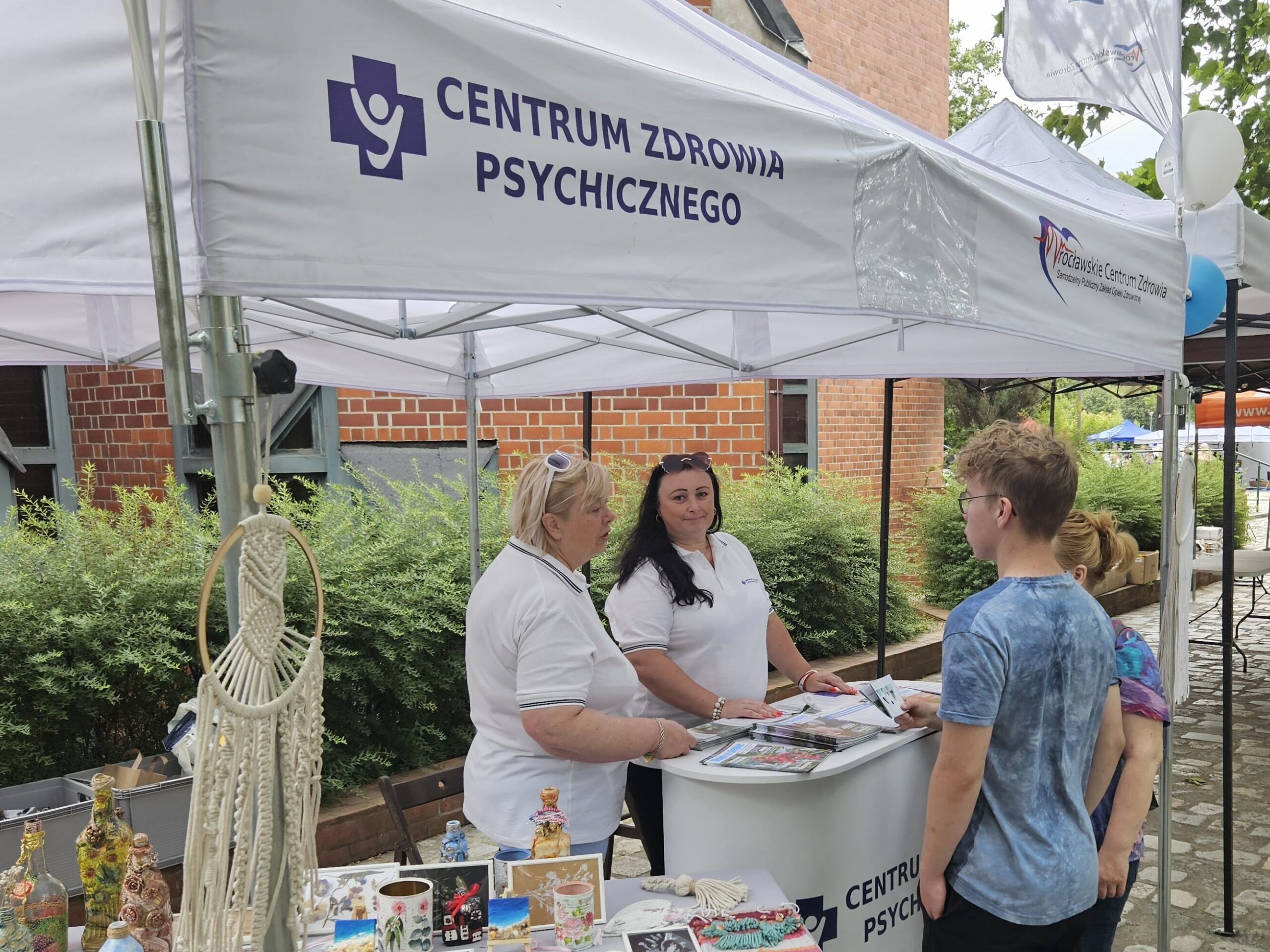 Dwie kobiety stoją za stołem z rękodziełem pod białym namiotem podczas wydarzenia plenerowego, rozmawiając z dwiema młodymi osobami.
