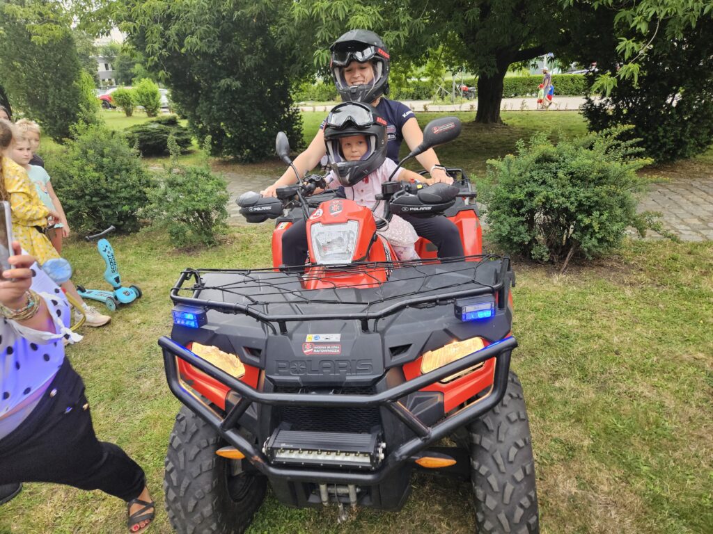 „Dziecko siedzi na quadzie ratowniczym obok dorosłego w kasku, a wokół stoją inni dorośli i dzieci podczas wydarzenia plenerowego.