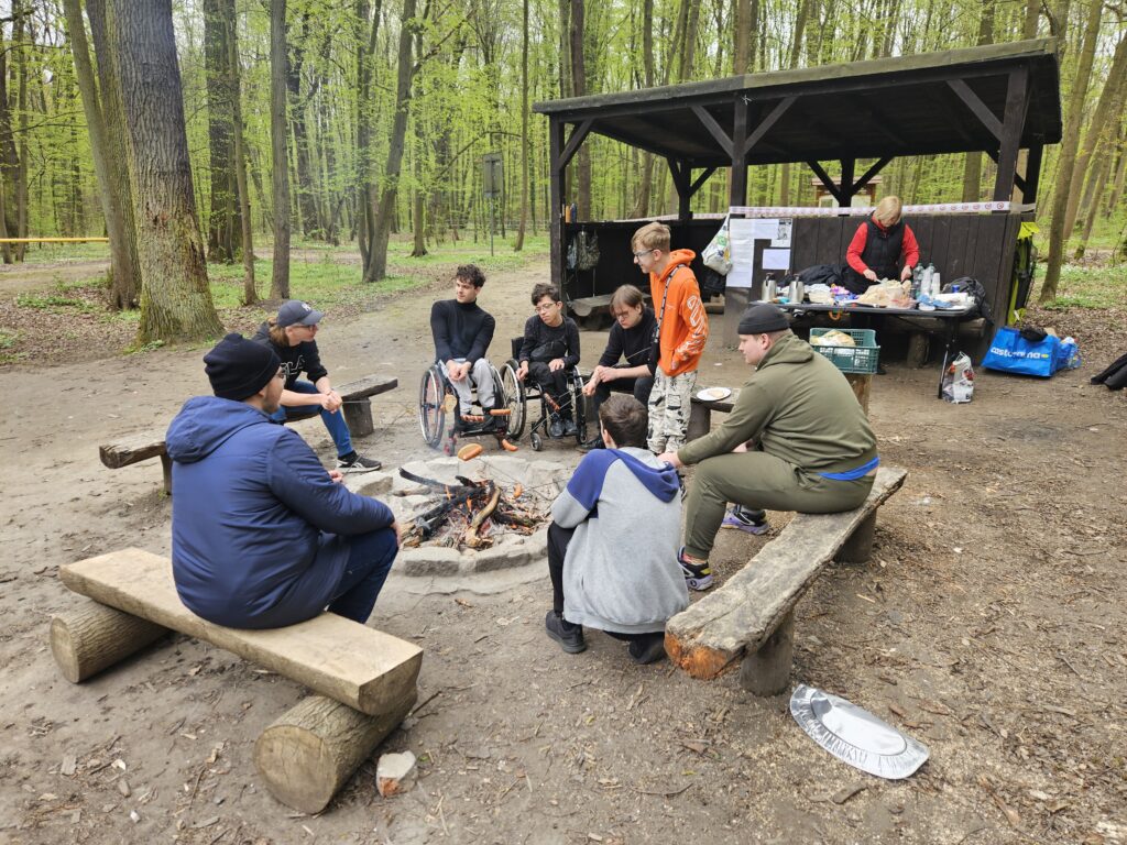 Grupa młodych osób, w tym dwie na wózkach, siedzi przy ognisku i piecze kiełbaski.