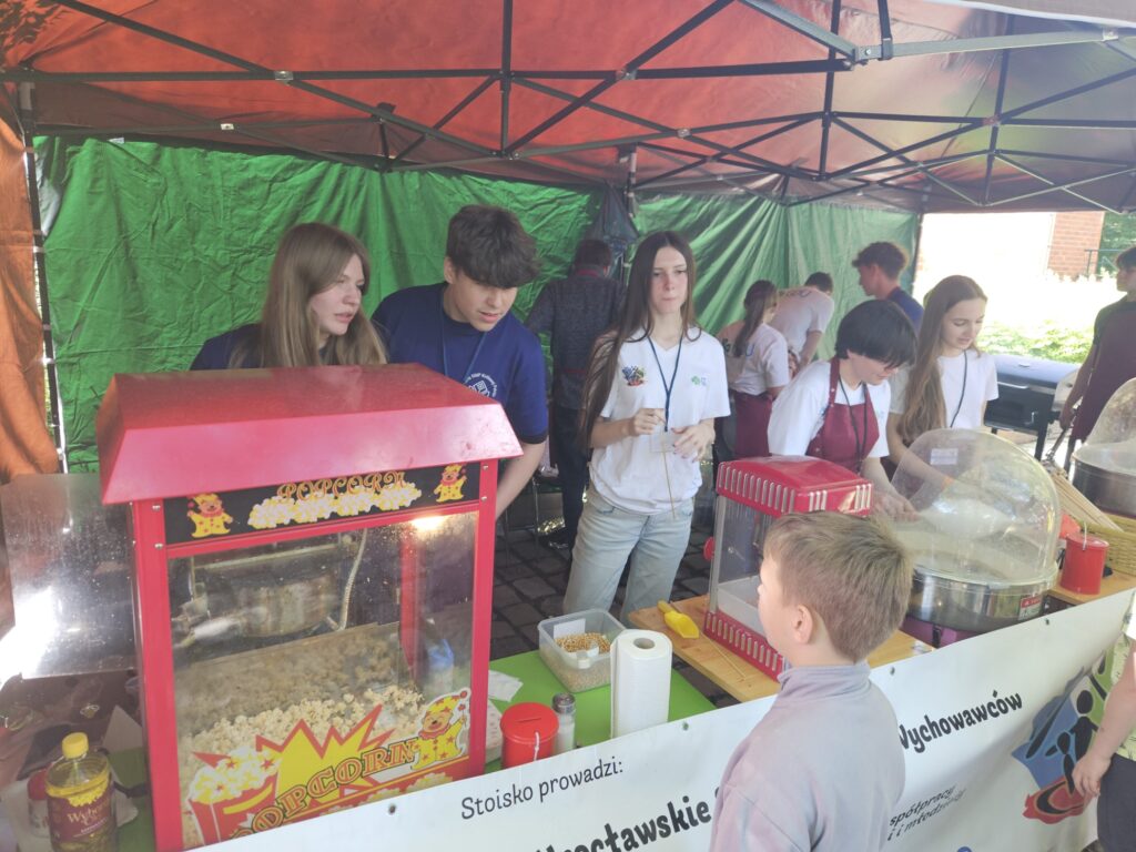 Kilka osób obsługuje stoisko z popcornem i watą cukrową pod czerwonym namiotem, a dziecko stoi przed ladą podczas wydarzenia na świeżym powietrzu.