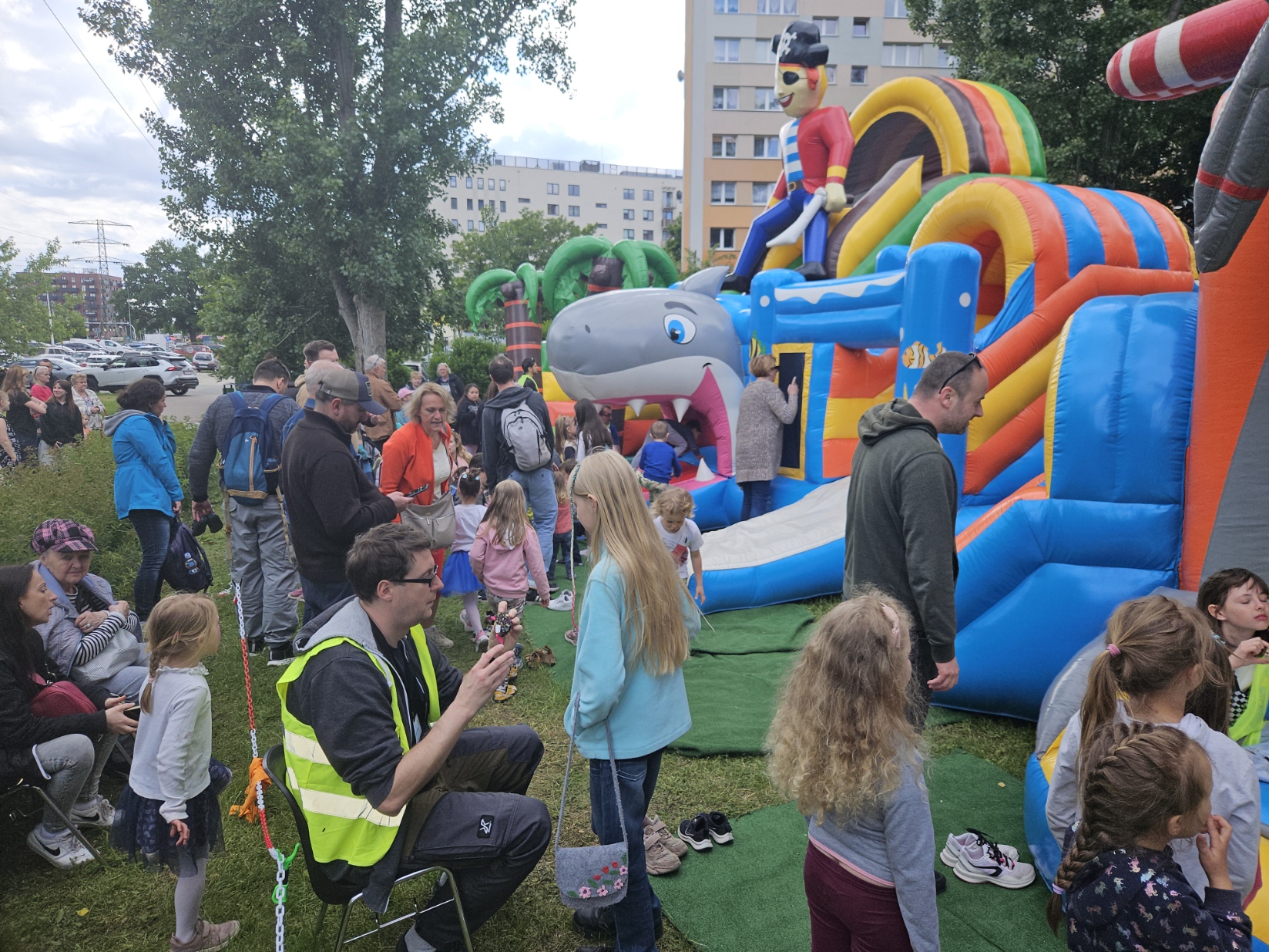 Dzieci bawią się przy kolorowej dmuchanej konstrukcji z wejściem w kształcie paszczy rekina, a dorośli stoją obok i obserwują podczas wydarzenia na świeżym powietrzu.
