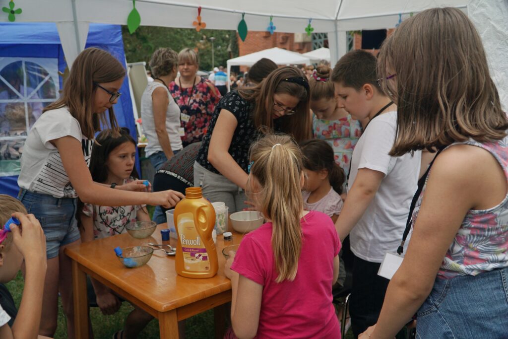 Dzieci i dorośli pracują przy stole z materiałami do doświadczeń pod białym namiotem podczas wydarzenia plenerowego.