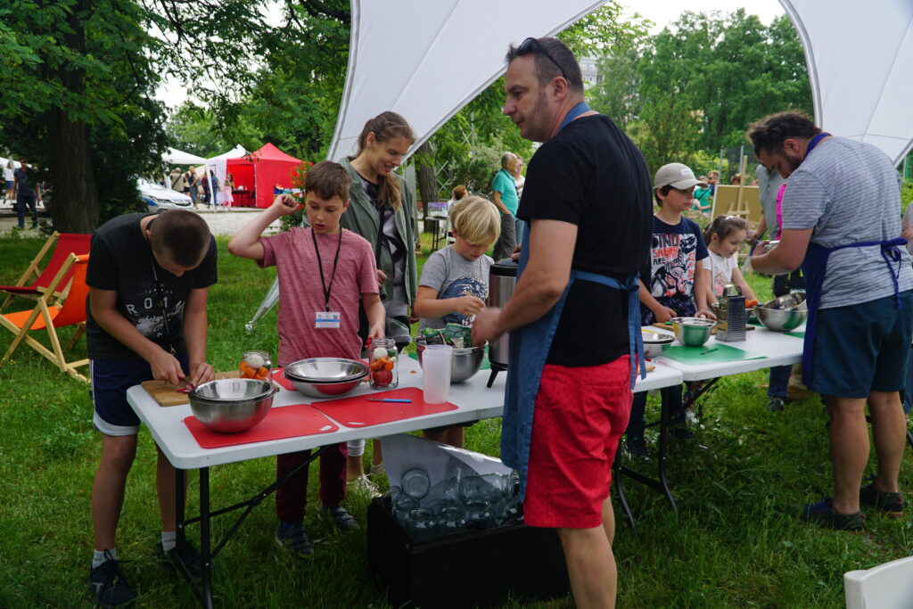 Dorośli i dzieci przygotowują jedzenie przy stołach pod namiotem podczas wydarzenia plenerowego.