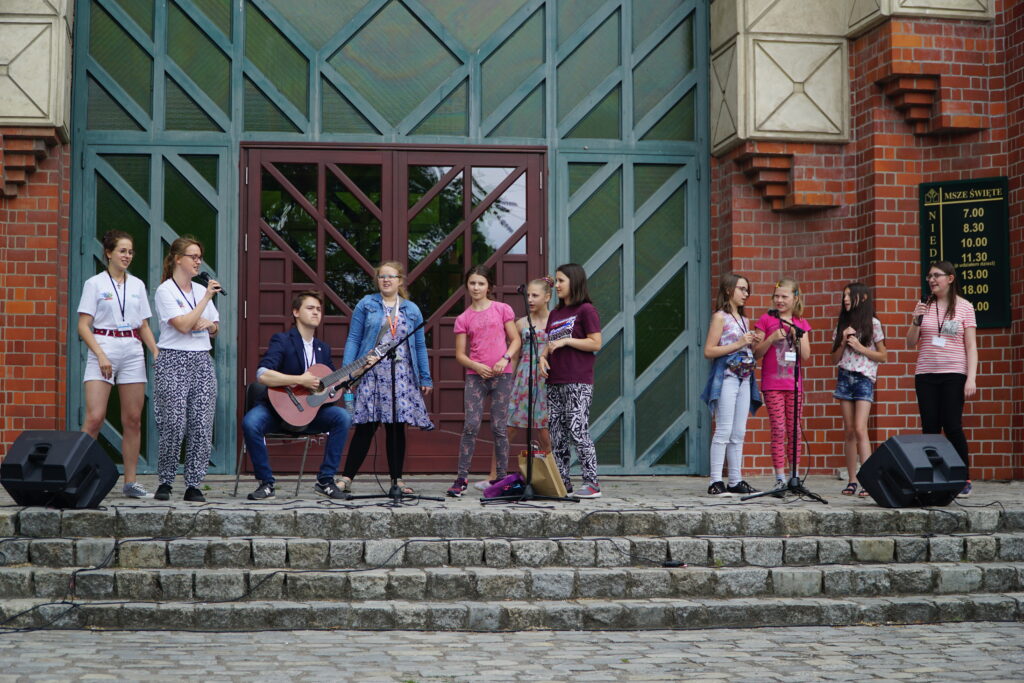 „Grupa dzieci i młodych osób stoi na kamiennej platformie przed budynkiem, a jedna osoba gra na gitarze podczas występu plenerowego.