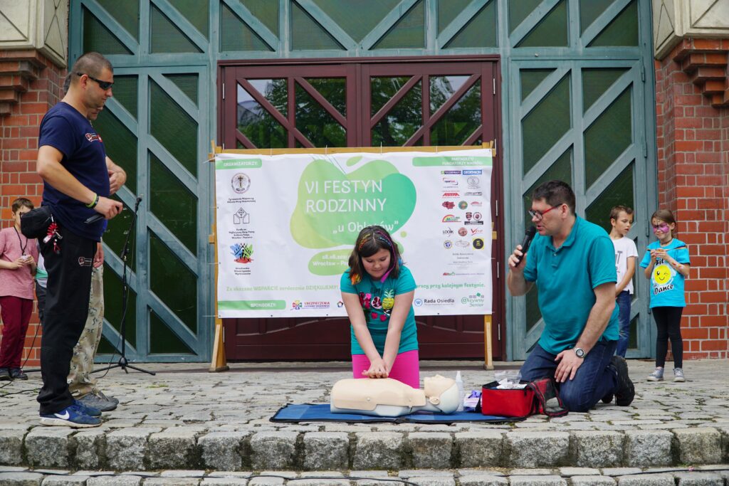 Dziewczynka ćwiczy uciskanie klatki piersiowej na fantomie, a obok niej klęczy mężczyzna mówiący do mikrofonu podczas pokazu pierwszej pomocy.