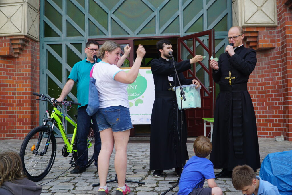 Dwaj mężczyźni w czarnych habitach prowadzą losowanie przed budynkiem, trzymając mikrofony i pudełko z karteczkami, a obok nich stoi mężczyzna z zielonym rowerem oraz kobieta unosząca ręce w geście radości.