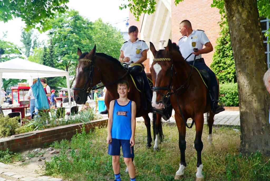 „Chłopiec stoi przed dwoma strażnikami miejskimi siedzącymi na brązowych koniach podczas wydarzenia plenerowego.